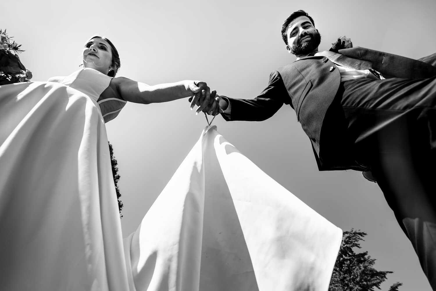 A candid black and white picture of a bride and groom holding hands, walking over a camera on their summer wedding day. 