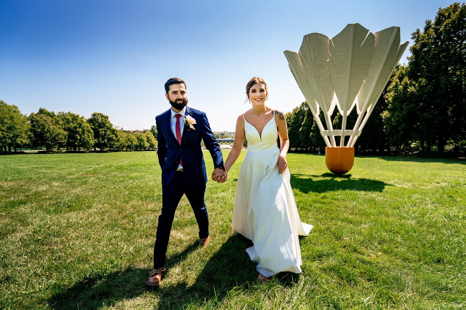 A colorful candid picture of a bride and groom holding hands, walking away from a giant shuttlecock on the lawn of the Nelson Atkins Museum of Art in Kansas City on their summer wedding day. 