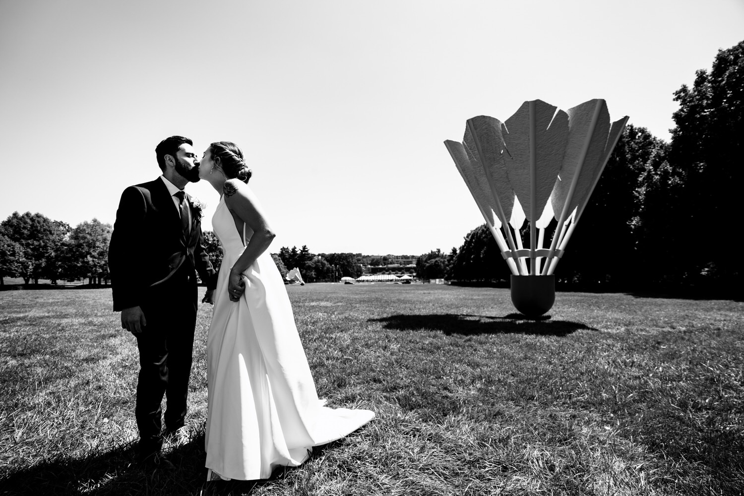 A candid black and white picture of a bride and groom leaning in to share a kiss, a giant shuttlecock visible on the grass behind them. 