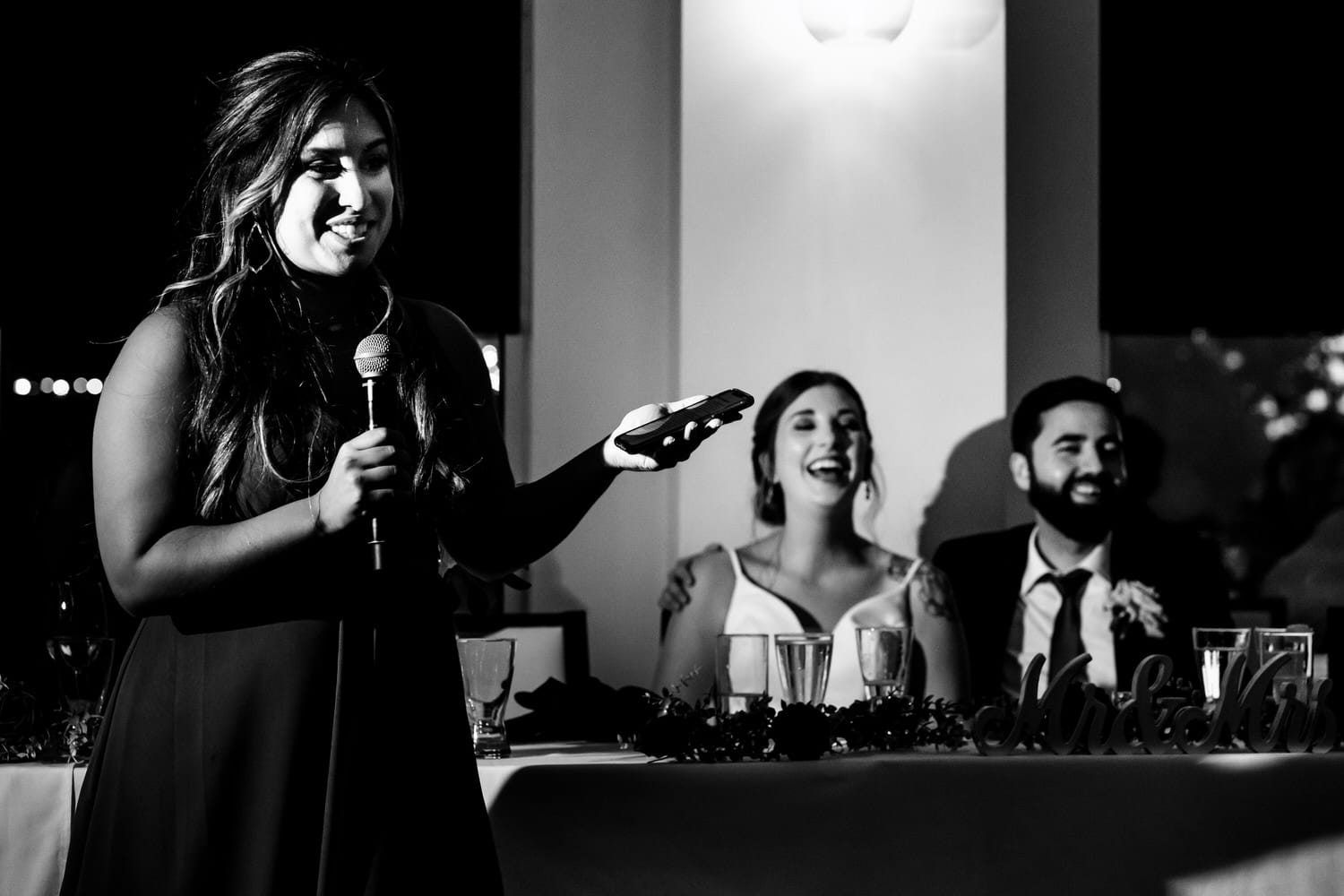 A candid black and white picture of a bridesmaid giving a toast, the bride and groom visible behind her during a classic reception at The Grand Street Cafe in Kansas City. 