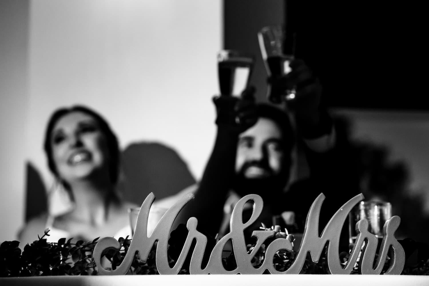 A candid black and white picture of a bride and groom raising their champagne flutes to toast, a wooden mr and mrs sign visible on the table in front of them during a classic wedding reception at The Grand Street Cafe in Kansas City. 