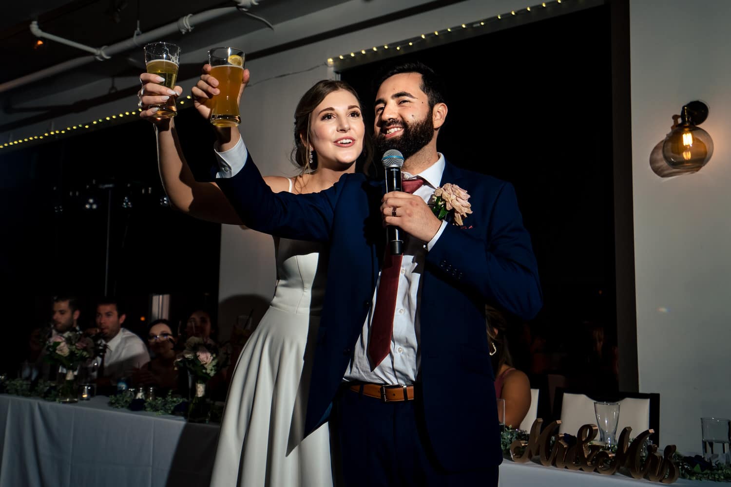 A colorful, candid picture of a bride and groom making a toast to their wedding guests, raising their beers in celebration during a classic wedding reception at The Grand Street Cafe in Kansas City. 