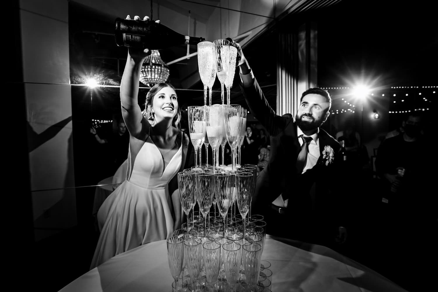 A candid black and white picture of a bride and groom pouring champagne into the very top of a champagne flute tower during a classic Kansas City wedding reception at The Grand Street Cafe. 