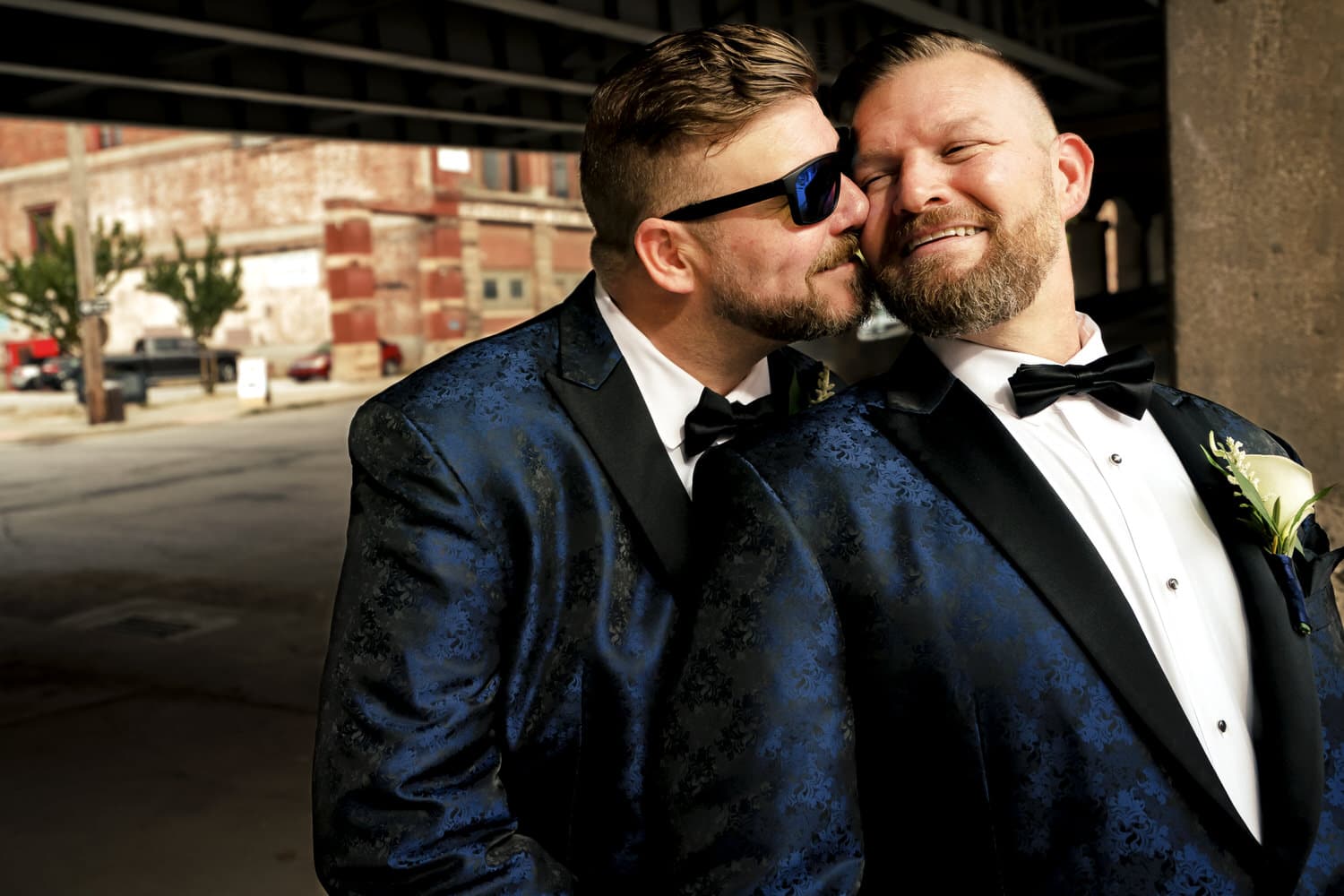 A candid, colorful, up-close picture of two grooms in navy blue tuxedos sharing an embrace on their fall wedding day in Kansas City. 