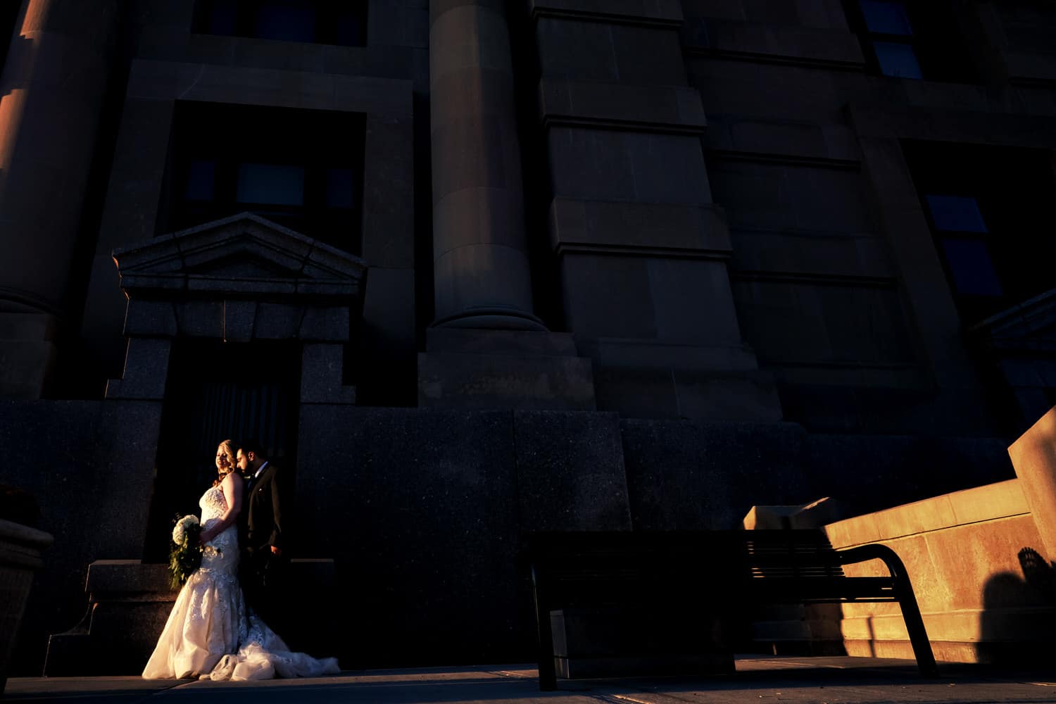A wide, dramatic portrait of a bride and groom sharing an embrace in a splash of sunlight in the front of Kansas City's Union Station on their fall wedding day. 