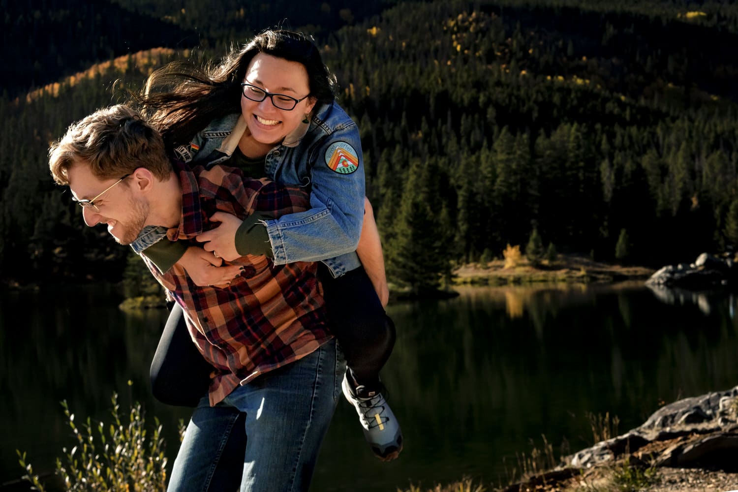 A candid, colorful picture of a woman in a jean jacket riding on the back of a man in a plaid shirty piggy-back, in front of a lake surrounded by evergreens and yellow aspen trees in Frisco, Colorado during an engagement session. 