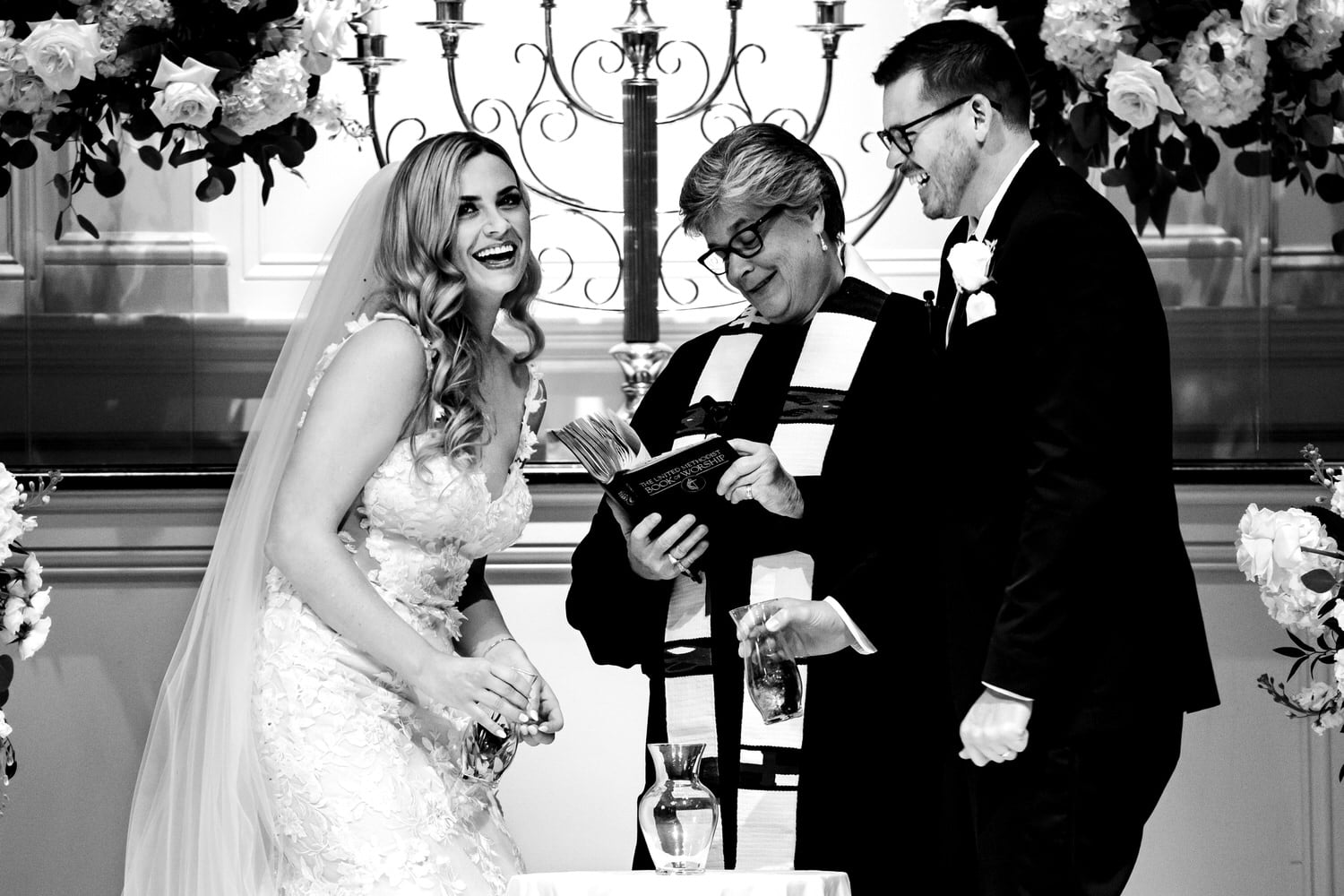 A candid black and white picture taken during a wedding ceremony of a bride in a long veil laughing and looking towards the camera as a groom in a black tuxedo laughs and looks at the bride as they get ready to pour sand into their unity jar during an black tie October wedding in Kansas City. 