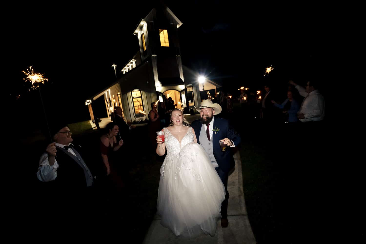 A candid picture of a bride holding a red drink and a groom wearing a cowboy hat running beneath two lines of sparklers, big smiles on their faces. 