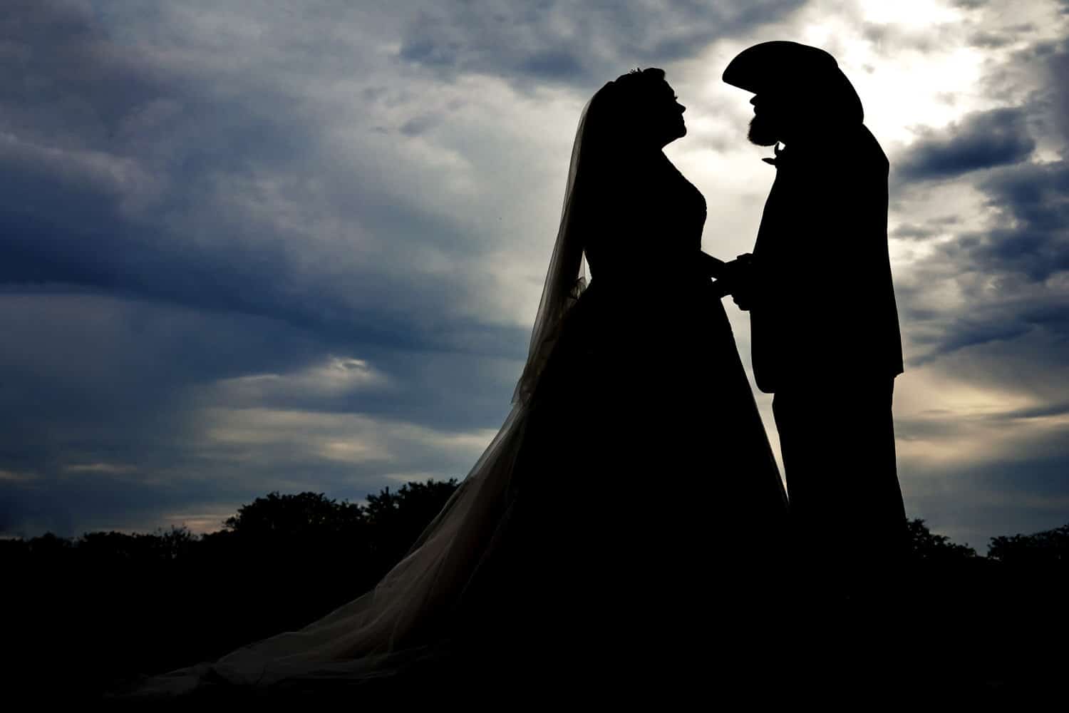 A woman in a wedding dress and a man in a cowboy hat hold hands, silhouetted in front of a stormy sky on their rustic fall wedding day at The Brim in Kansas City. 