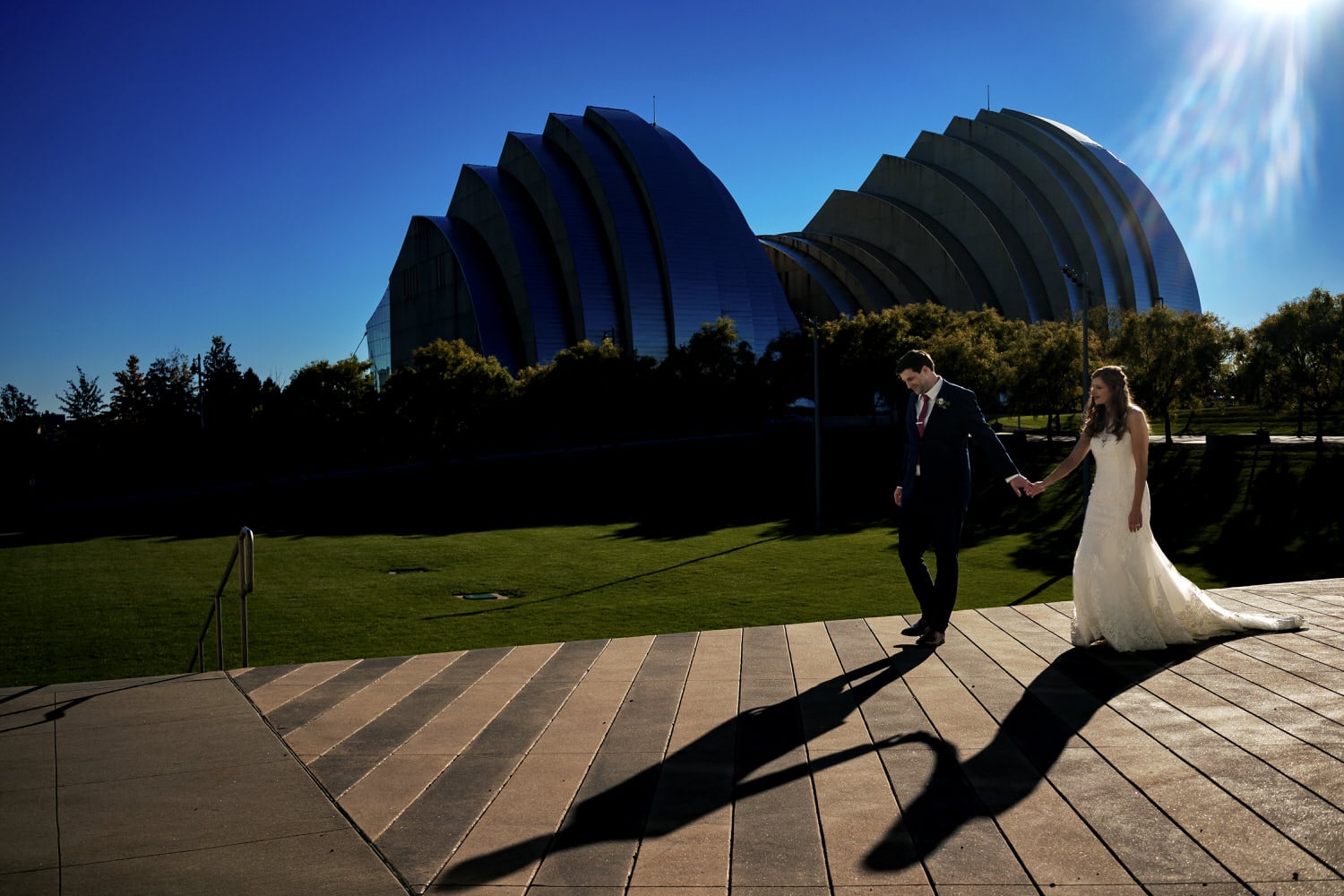 A colorful, candid picture of a bride and groom holding hands and walking across a black and white striped pavement, the Kauffman Center for the Performing Arts visible in the background on a fall wedding day in Kansas City. 