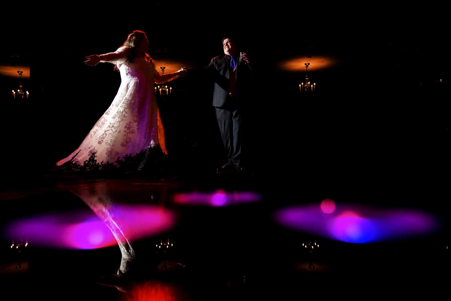 A candid colorful picture of a man in a black tuxedo twirling a woman in a white wedding dress with black lace across the dance floor during their Halloween wedding reception in Kansas City. 