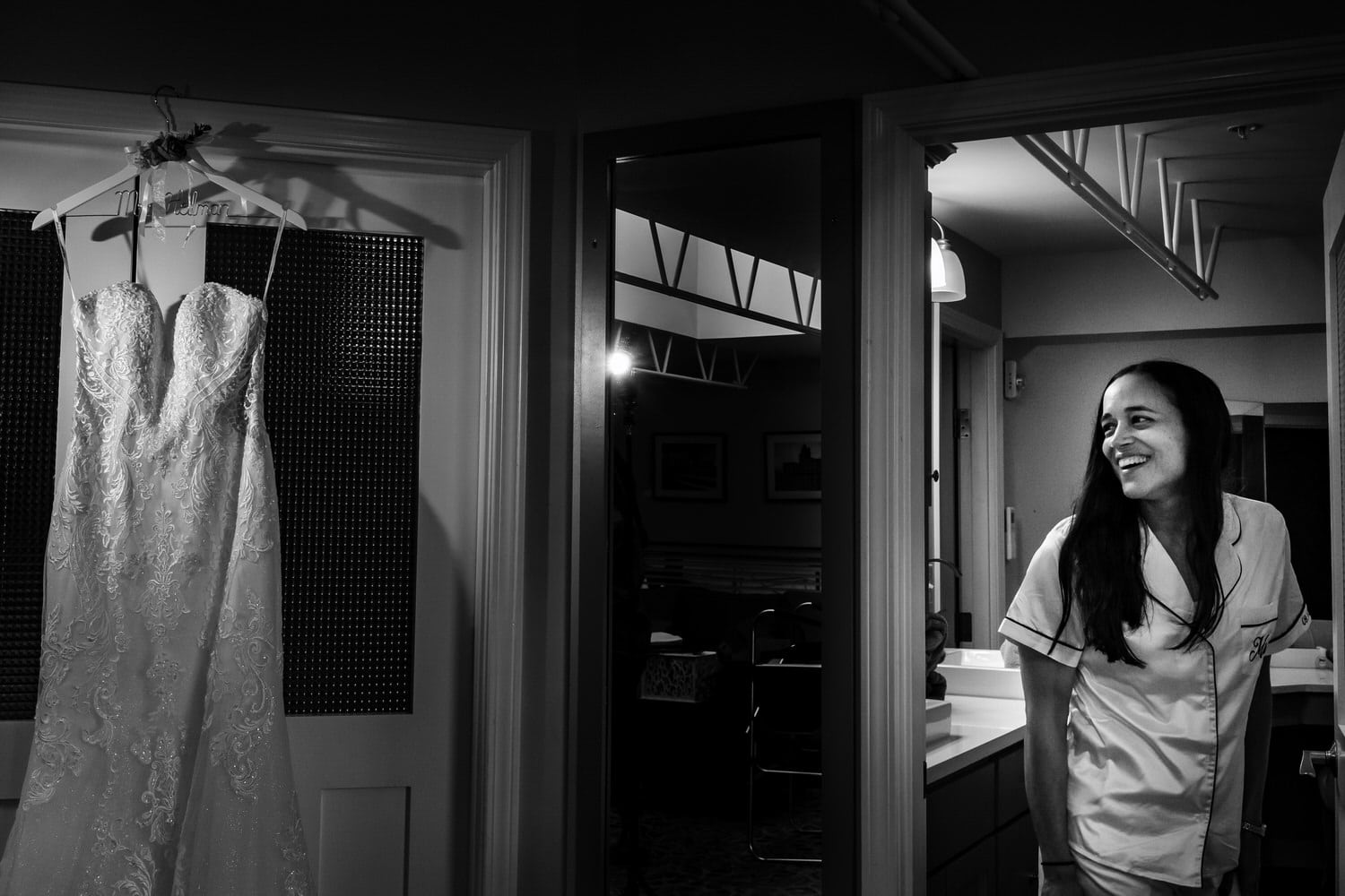 A candid black and white picture of a woman in white pajamas peeking around a door corner to see her wedding dress hanging in a doorway on her wedding day. 