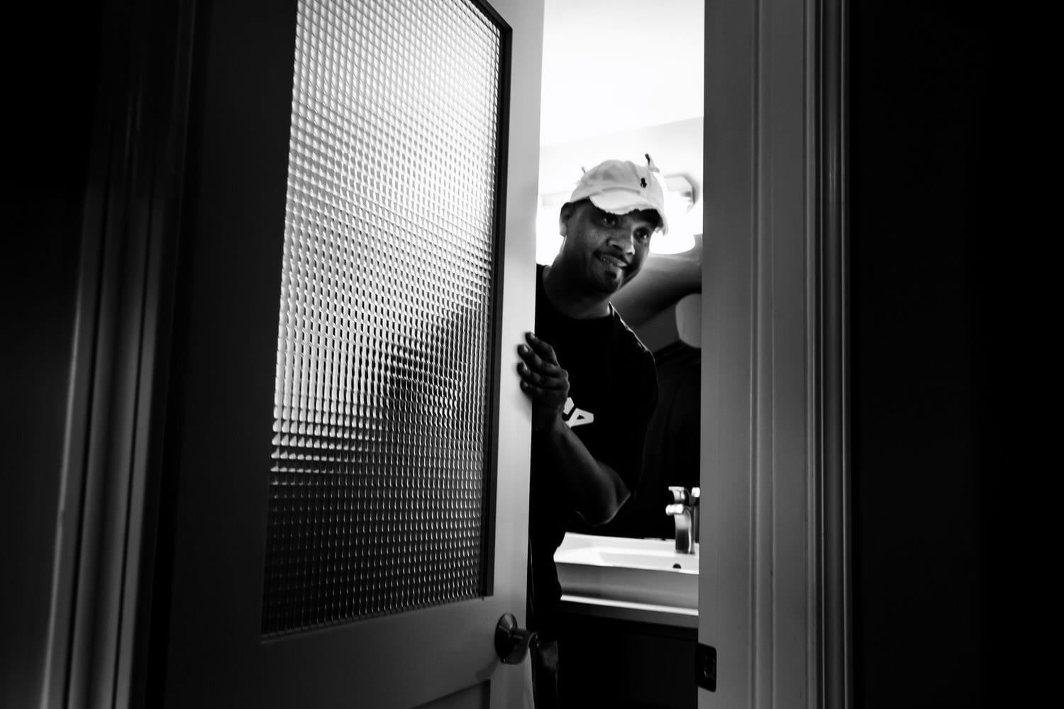A candid black and white picture of a man peeking through a partially  open door, a smile on his face. 