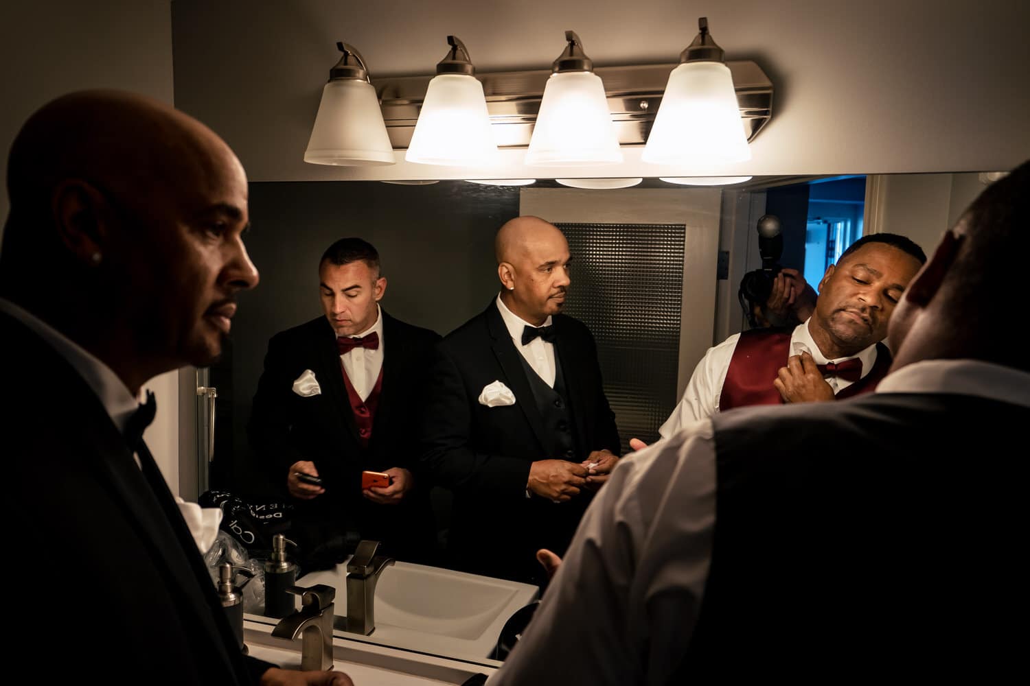 A candid picture of a group of men getting ready for a wedding in front of a large bathroom mirror. 