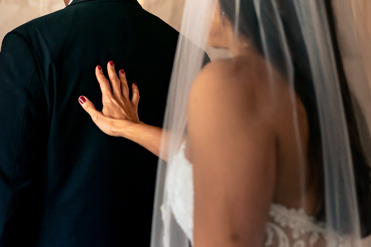A close-up picture of a bride reaching out to tap her groom on the shoulder, her red fingernails visible on his back on their summer wedding day at 28 Event Space in Kansas City. 