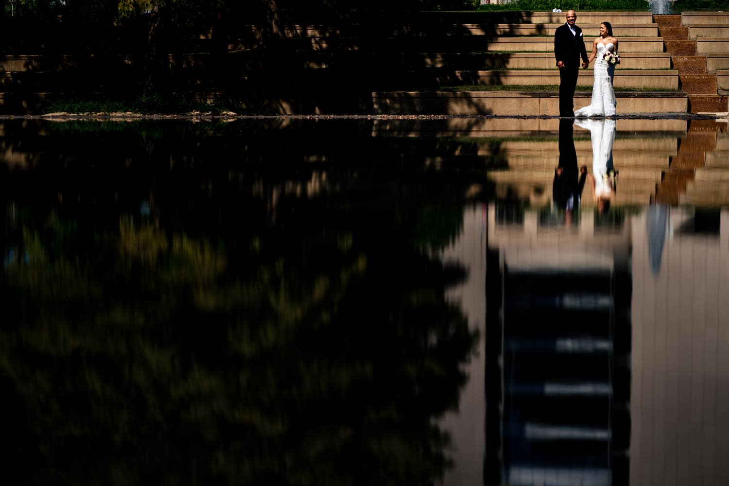 A wide, colorful portrait of a bride and groom holding hands, walking in front of a reflective pool of water on their summer wedding day in Kansas City. 