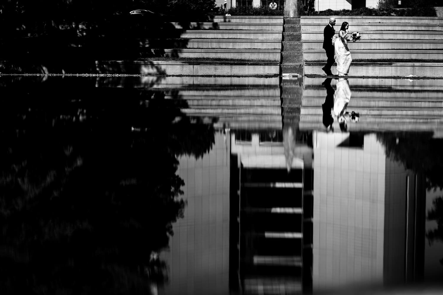 A candid black and white picture of a bride and groom walking in front of a reflective pool of water on their summer wedding day in Kansas City. 