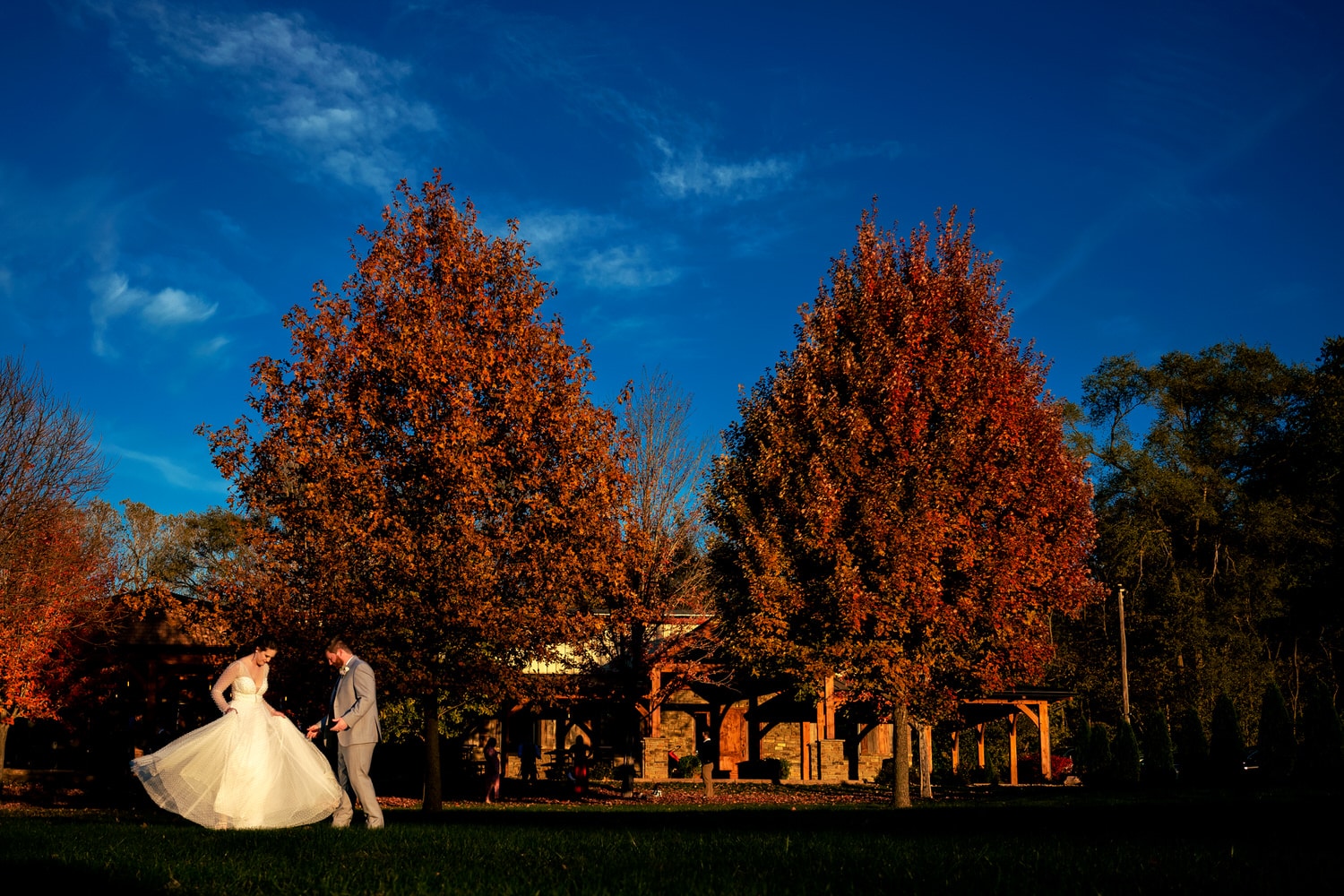A colorful, candid picture of a bride twirling her wedding dress as her groom looks on in front of colorful, red trees on their fall wedding day in Kansas City. 