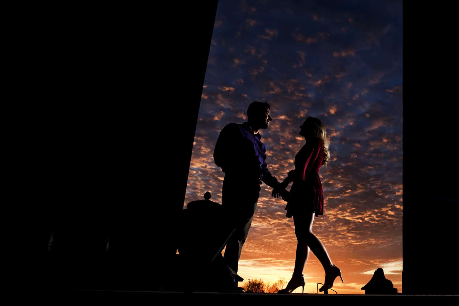 A colorful, candid picture of a vibrant sunset, a woman in high heels and a red dress walking towards a man in a purple shirt during an engagement session at Liberty Memorial in Kansas City. 