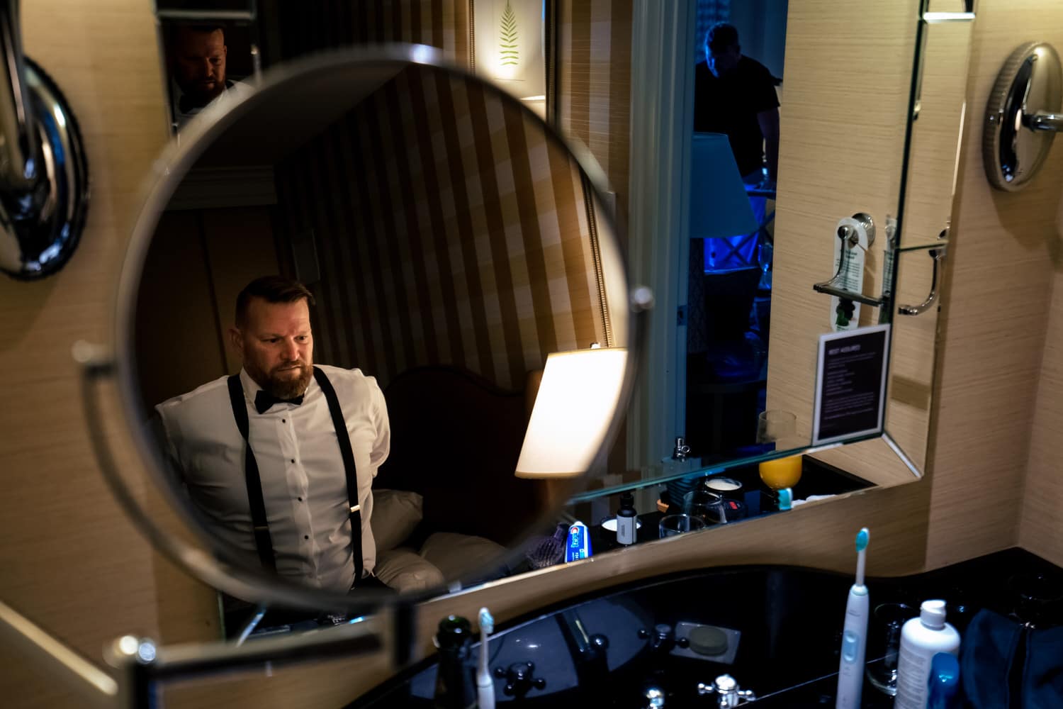 A candid picture taken in the reflection of a mirror of a groom in suspenders tucking his dress shirt into his pants. 