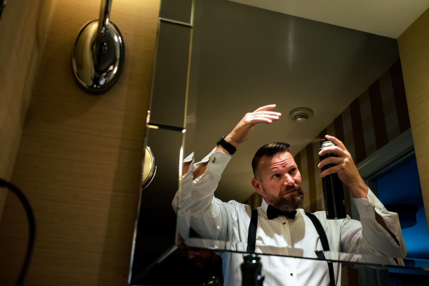A candid picture of a groom spraying his hair with hair spray on the morning of his fall wedding. 