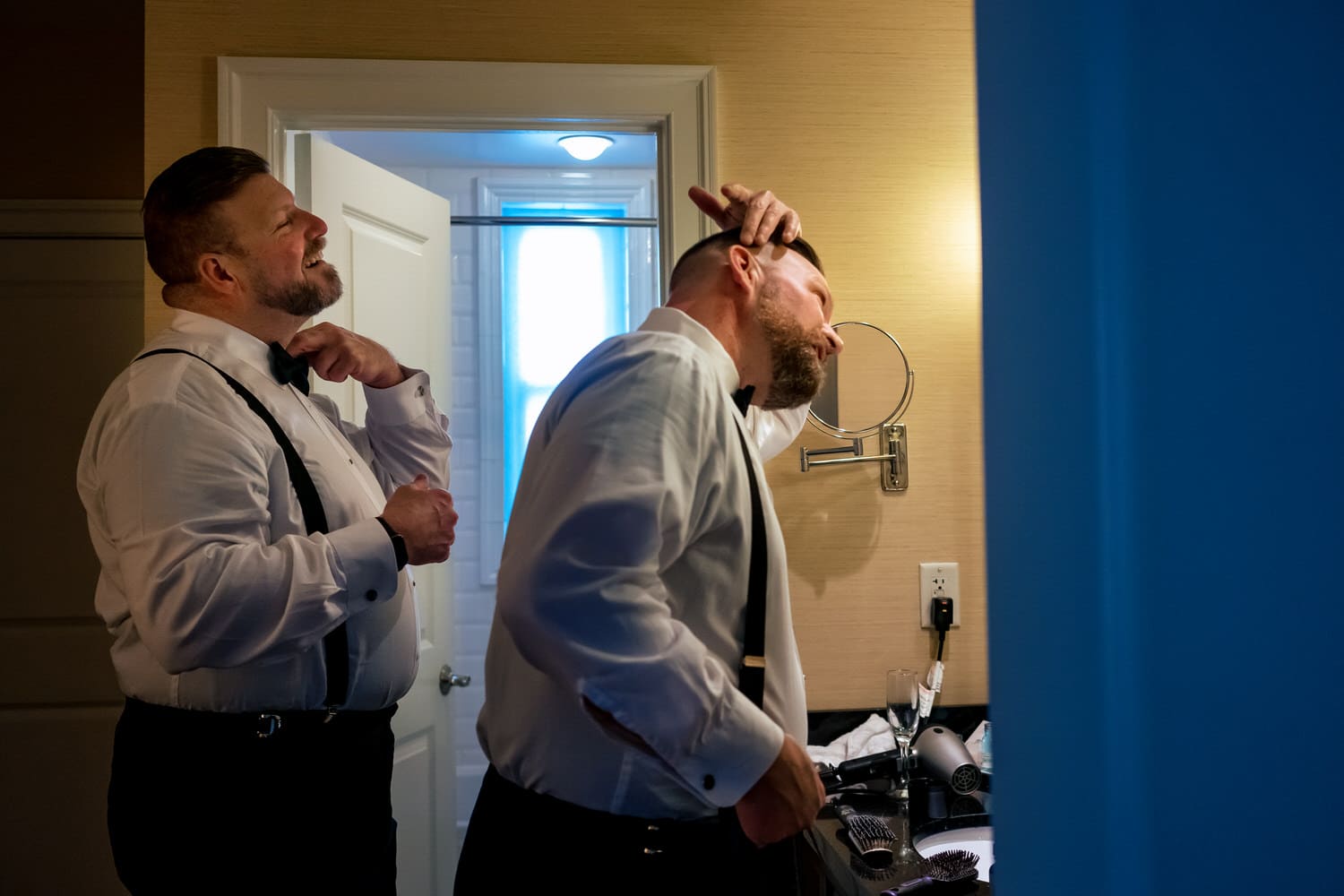 A candid picture of two grooms adjusting their hair and bow ties while looking in a mirror on the morning of their wedding. 