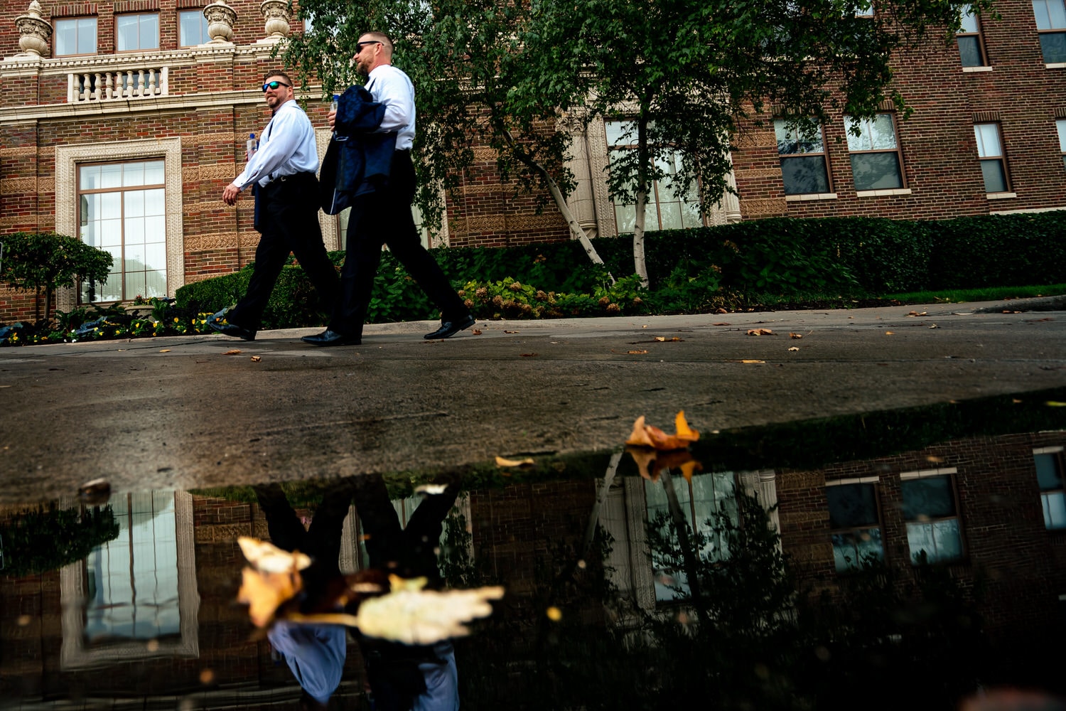 A candid picture of two grooms in tuxedos walking on a sidewalk, their reflections visible in a puddle in front of them. 