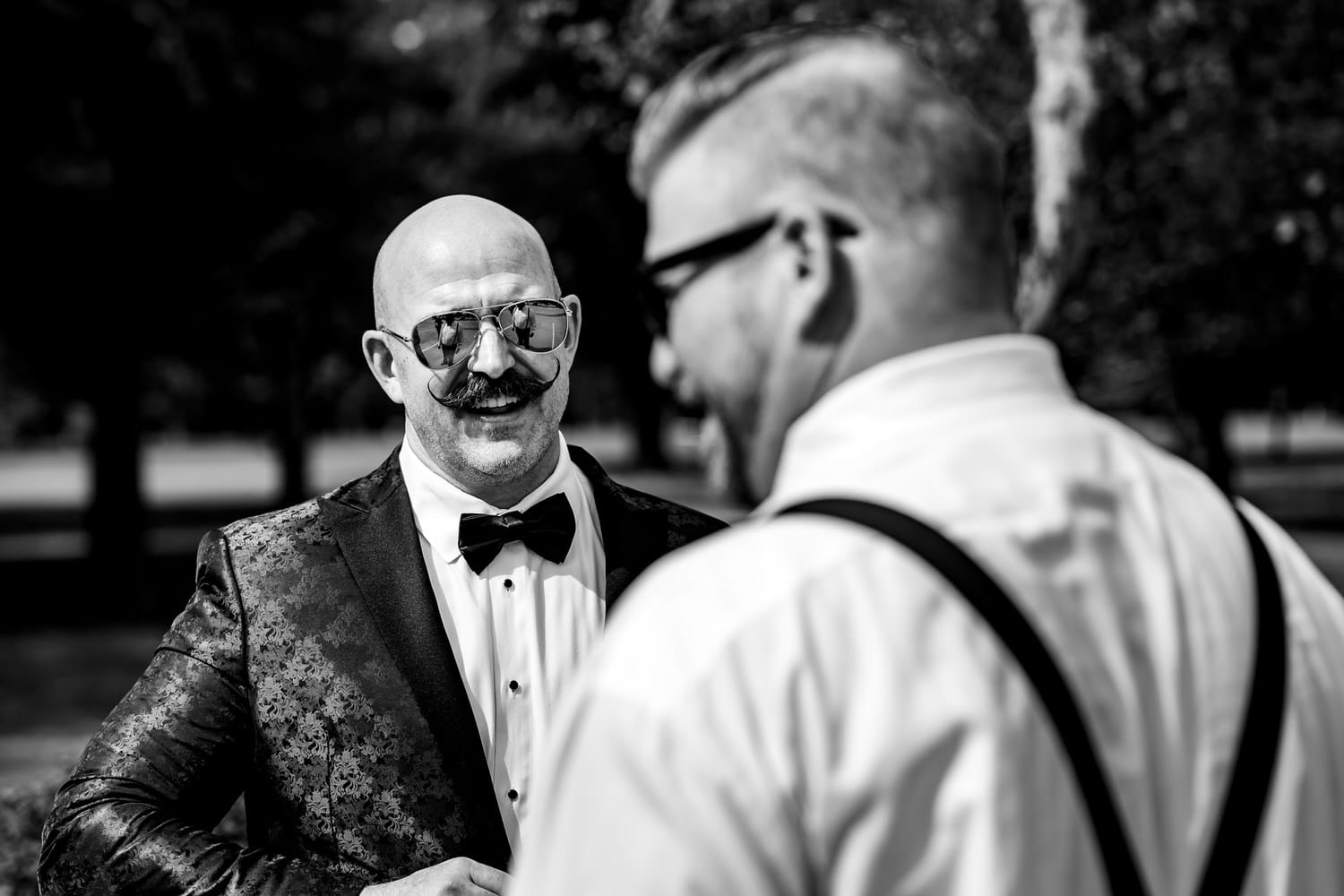 A candid black and white picture of a bald groomsman in sunglasses smiling at a groom. 