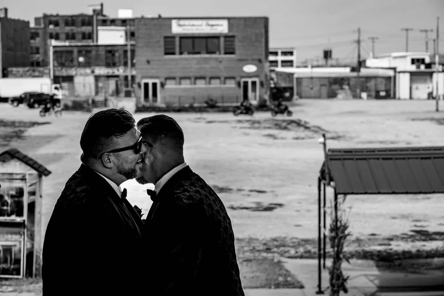 A candid black and white picture of a groom laughing and whispering into his groom's ear, Kansas City's West Bottoms neighborhood visible behind them. 
