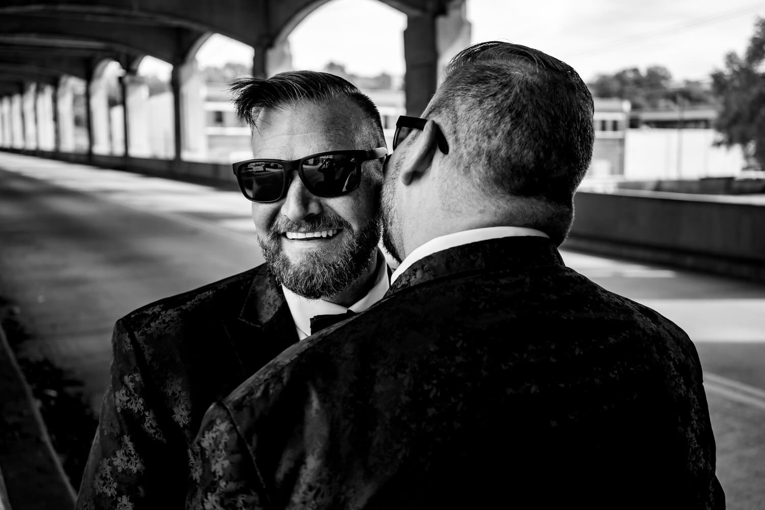 A candid black and white picture of a groom laughing and whispering into his groom's ear, Kansas City's West Bottoms neighborhood visible behind them. 