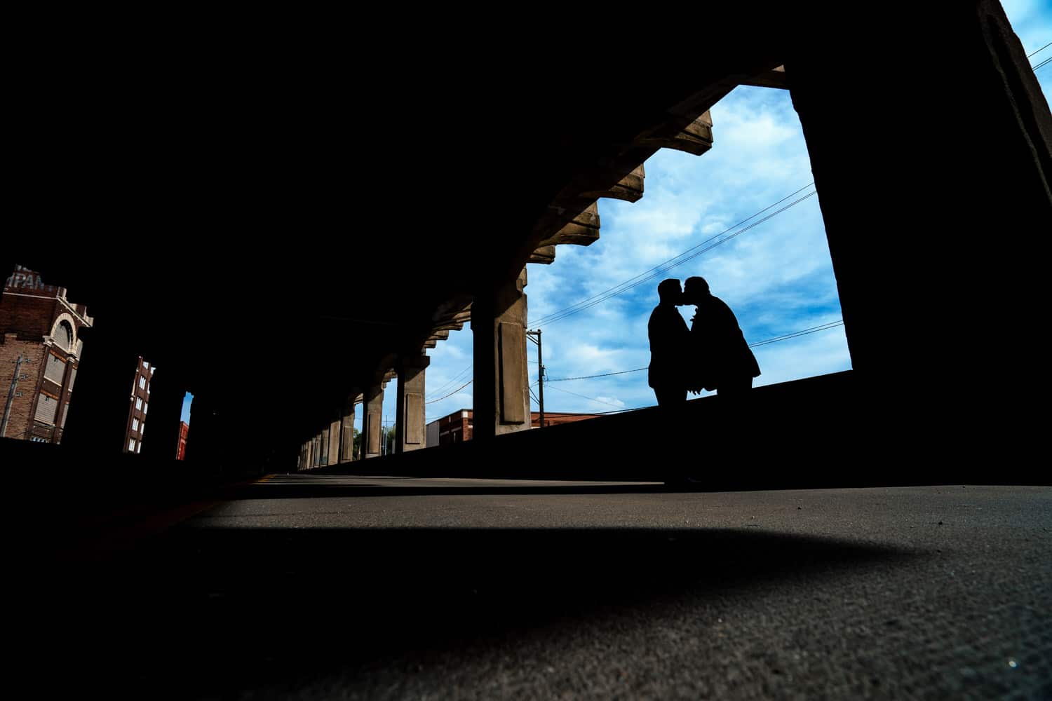 A colorful portrait of two grooms silhouetted in an opening of the 12th Street Viaduct, blue sky visible behind them on their fall wedding day in Kansas City. 