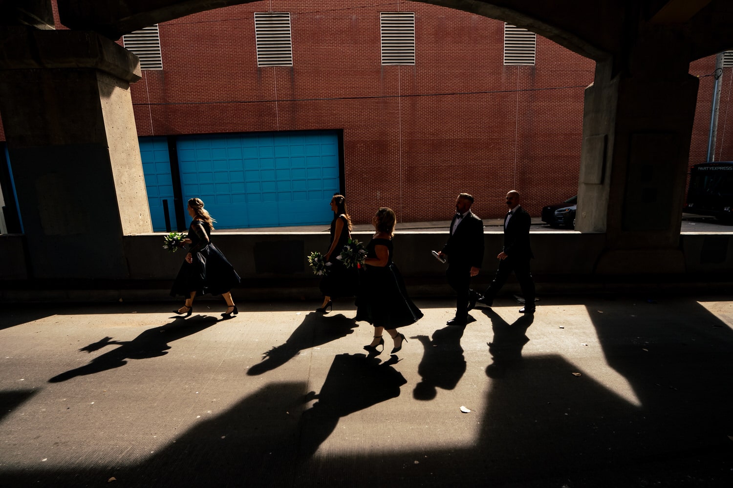 A colorful, semi-silhouetted picture of a wedding party walking through a sunny spot on the 12th Street Viaduct, their shadows visible in front of them on a sunny fall wedding day. 