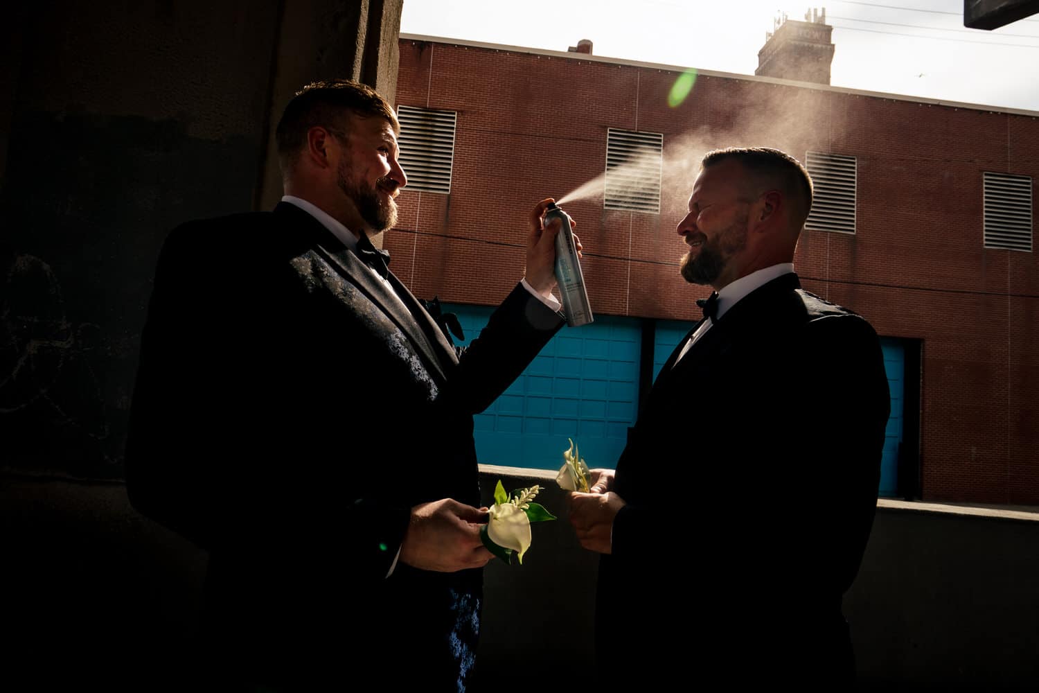 A candid, colorful picture of a groom spraying his partner with hairspray; the hairspray backlight by the sun on a fall wedding day in Kansas City. 