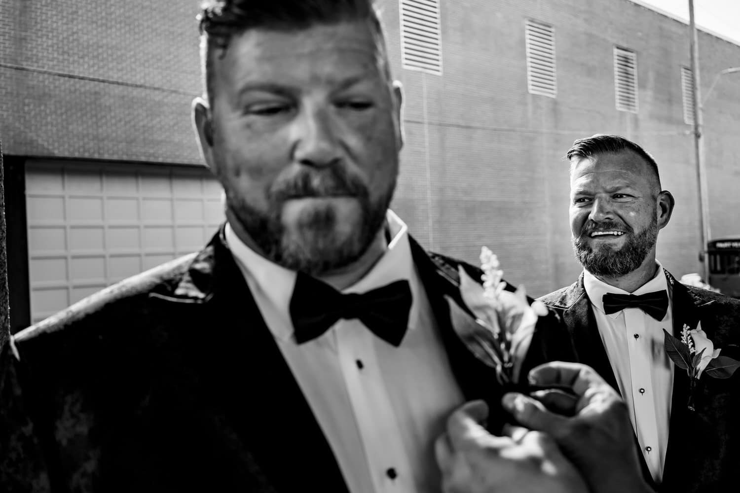 A candid black and white picture of a groom getting a boutionniere pinned on his jacket as his groom watches on from the background. 