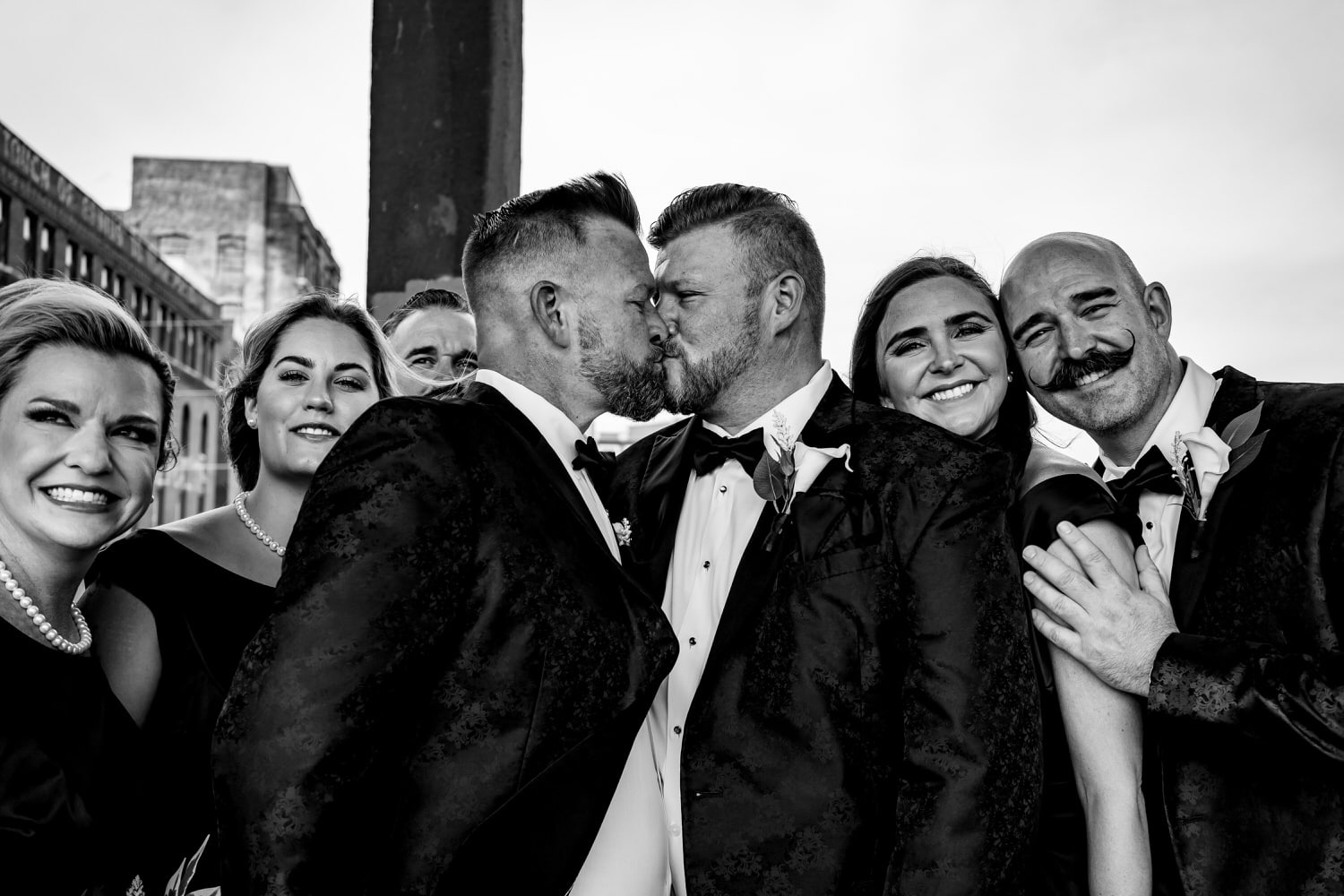 A candid black and white picture of two grooms sharing a kiss surrounded by their wedding party on a fall wedding day in Kansas City. 