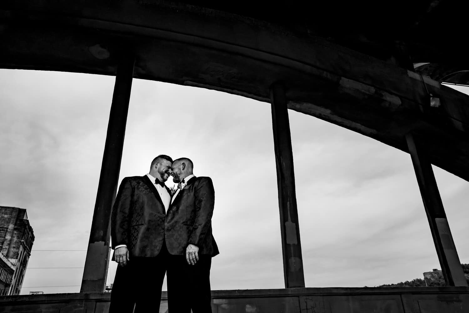 A candid black and white picture of two grooms leaning in to share an embrace on the 12th Street Viaduct on their fall wedding day in Kansas City. 