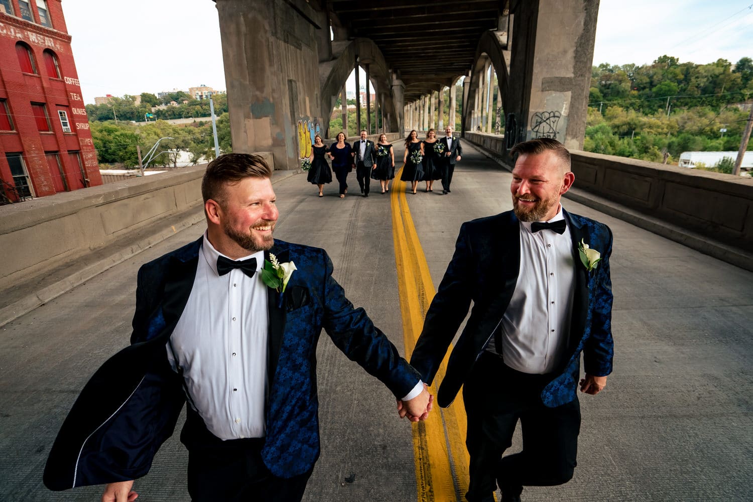 A candid picture of two grooms walking down the 12th Street Viaduct holding hands, looking at each other and laughing as their wedding party walks behind them on a fall wedding day in Kansas City. 