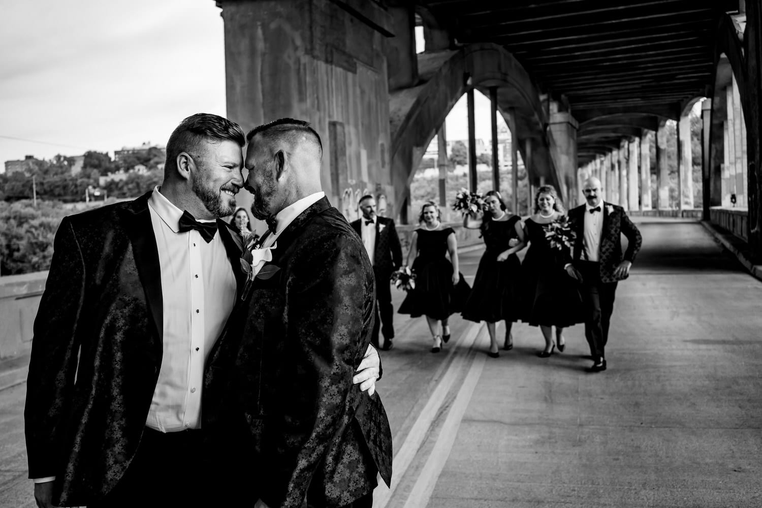 A candid picture of two grooms walking down the 12th Street Viaduct holding hands, looking at each other and laughing as their wedding party walks behind them on a fall wedding day in Kansas City. 