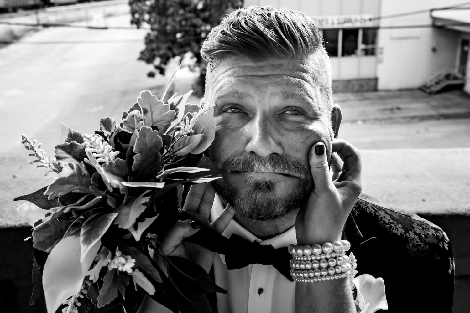 A candid black and white picture of a groom with a mustache getting his cheeks pinched by a woman with a pearl bracelet and bouquet on a fall wedding day in Kansas City. 