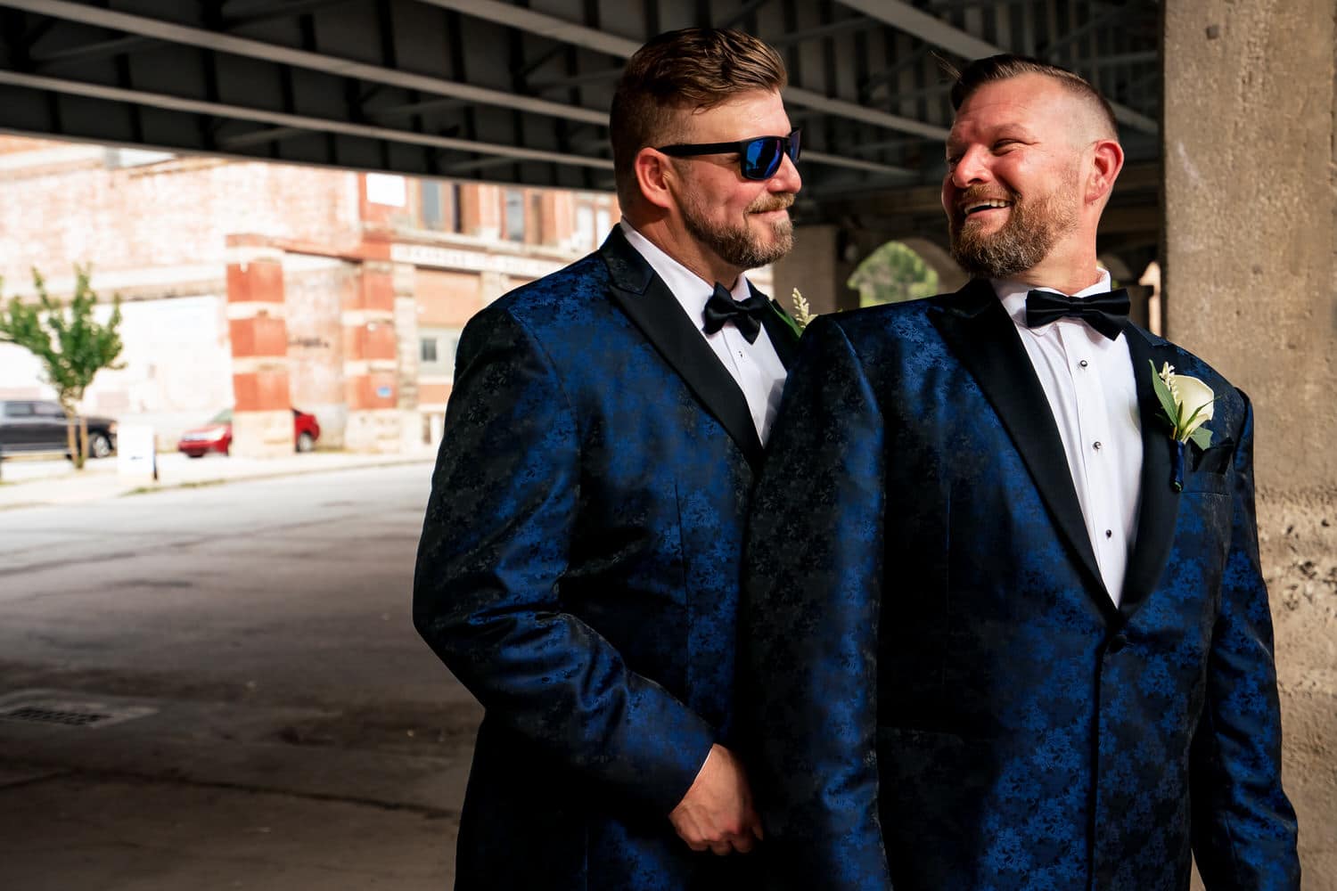 A colorful portrait of two grooms in blue tuxedos sharing an embrace and laughing together in Kansas City's West Bottoms on their fall wedding day. 