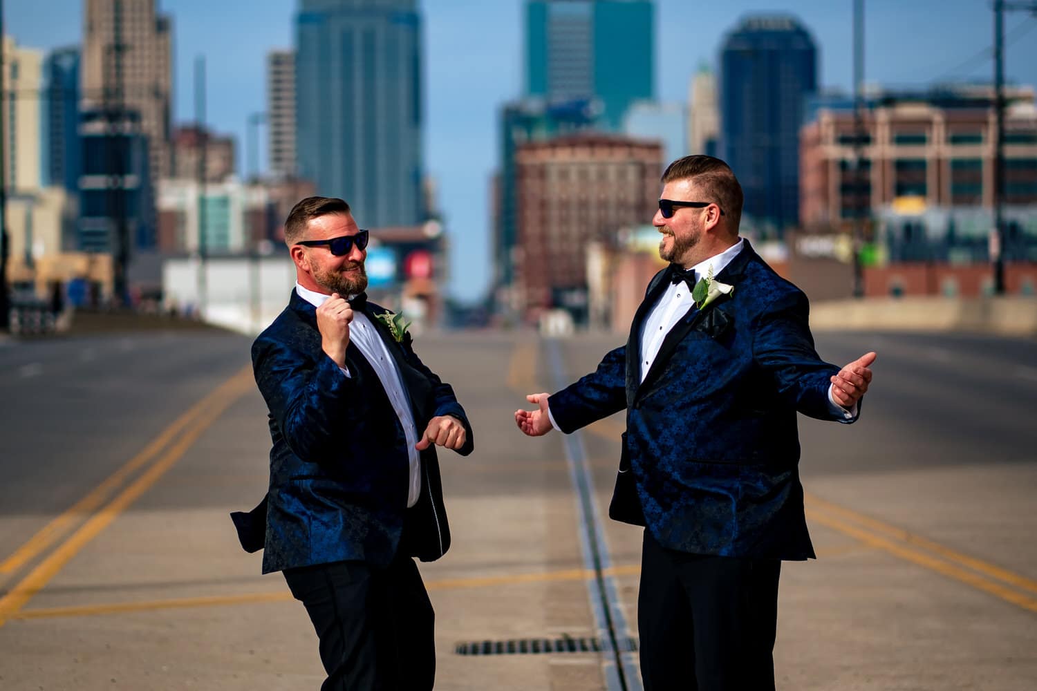 A colorful, candid picture of two grooms holding hands, walking down the center of Main Street, the Kansas City skyline visible behind them on a fall wedding day in Kansas City. 