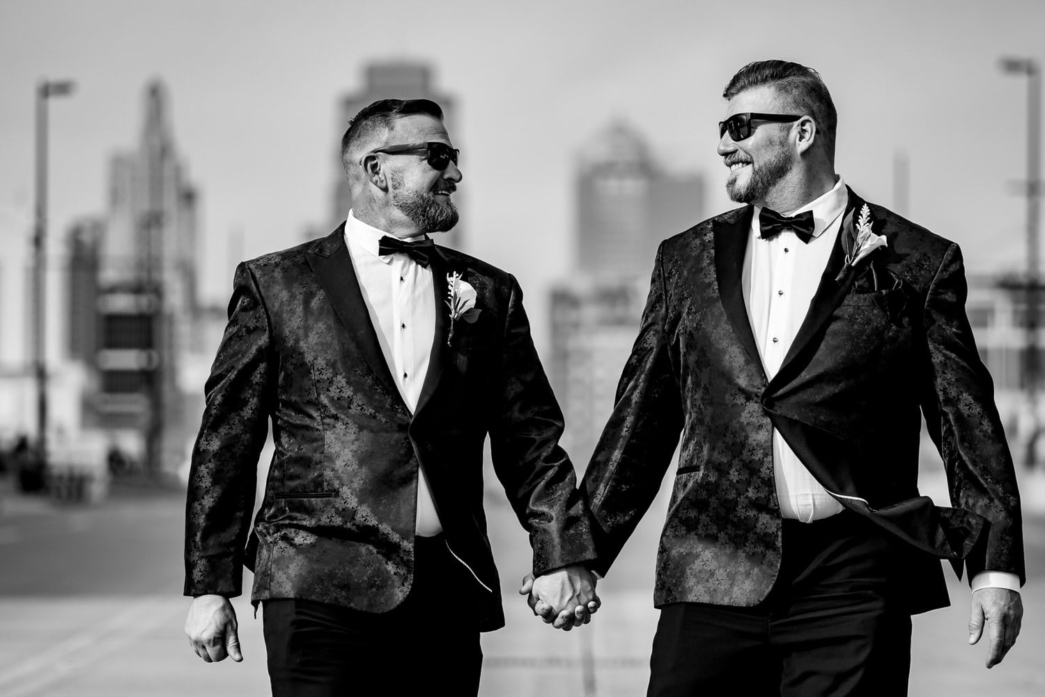 A colorful, candid picture of two grooms holding hands, walking down the center of Main Street, the Kansas City skyline visible behind them on a fall wedding day in Kansas City. 