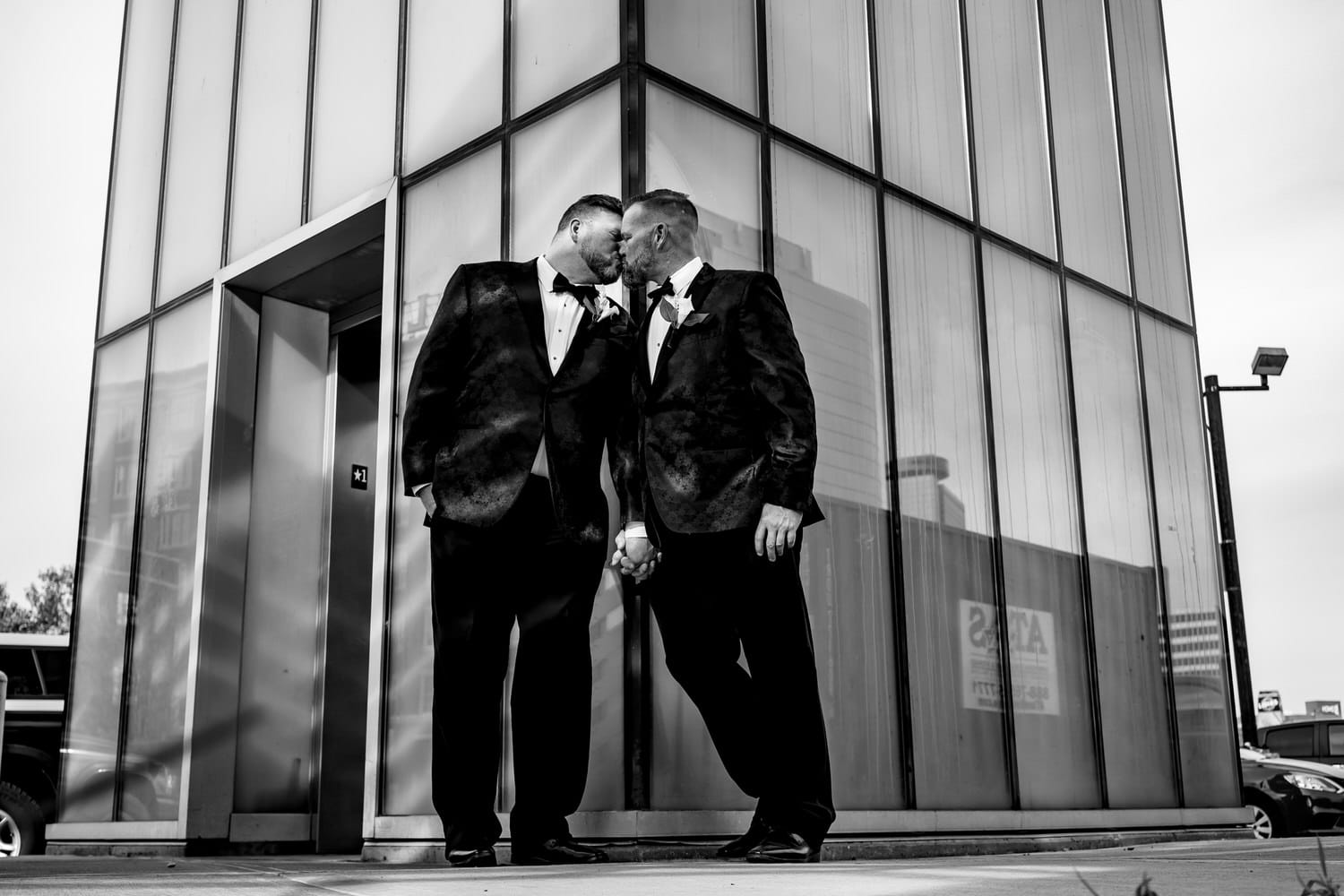 A black and white picture of two grooms leaning up against a modern building, sharing a kiss on their fall wedding day in Kansas City. 