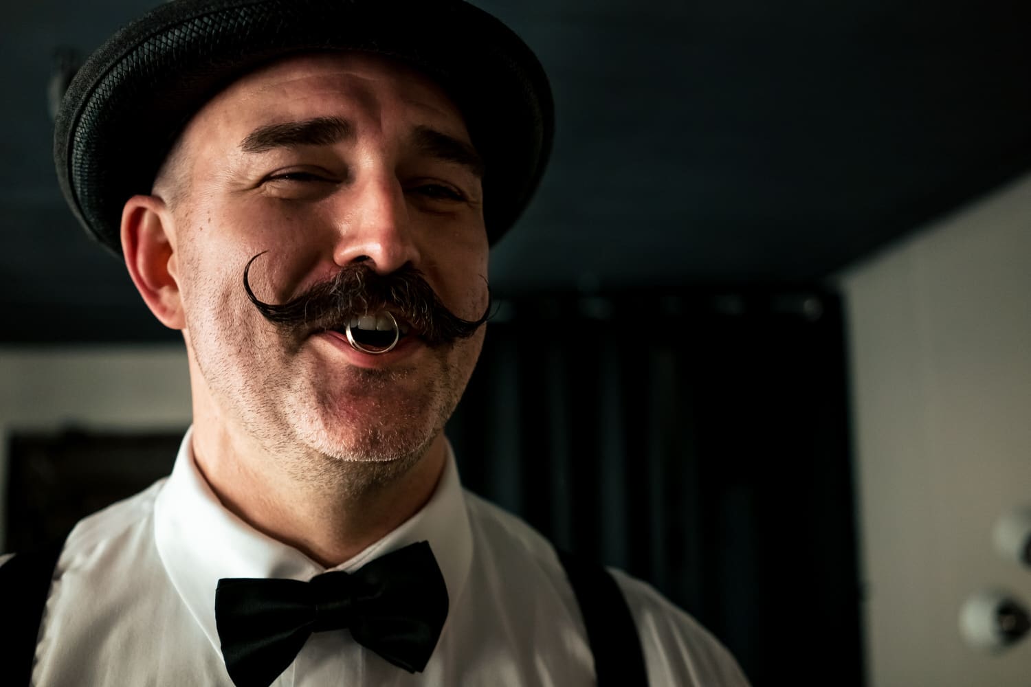 A candid picture of a groomsman with a handlebar mustache holding a silver ring in between his lips, smiling at the camera on a fall wedding day at The Madrid Theatre. 