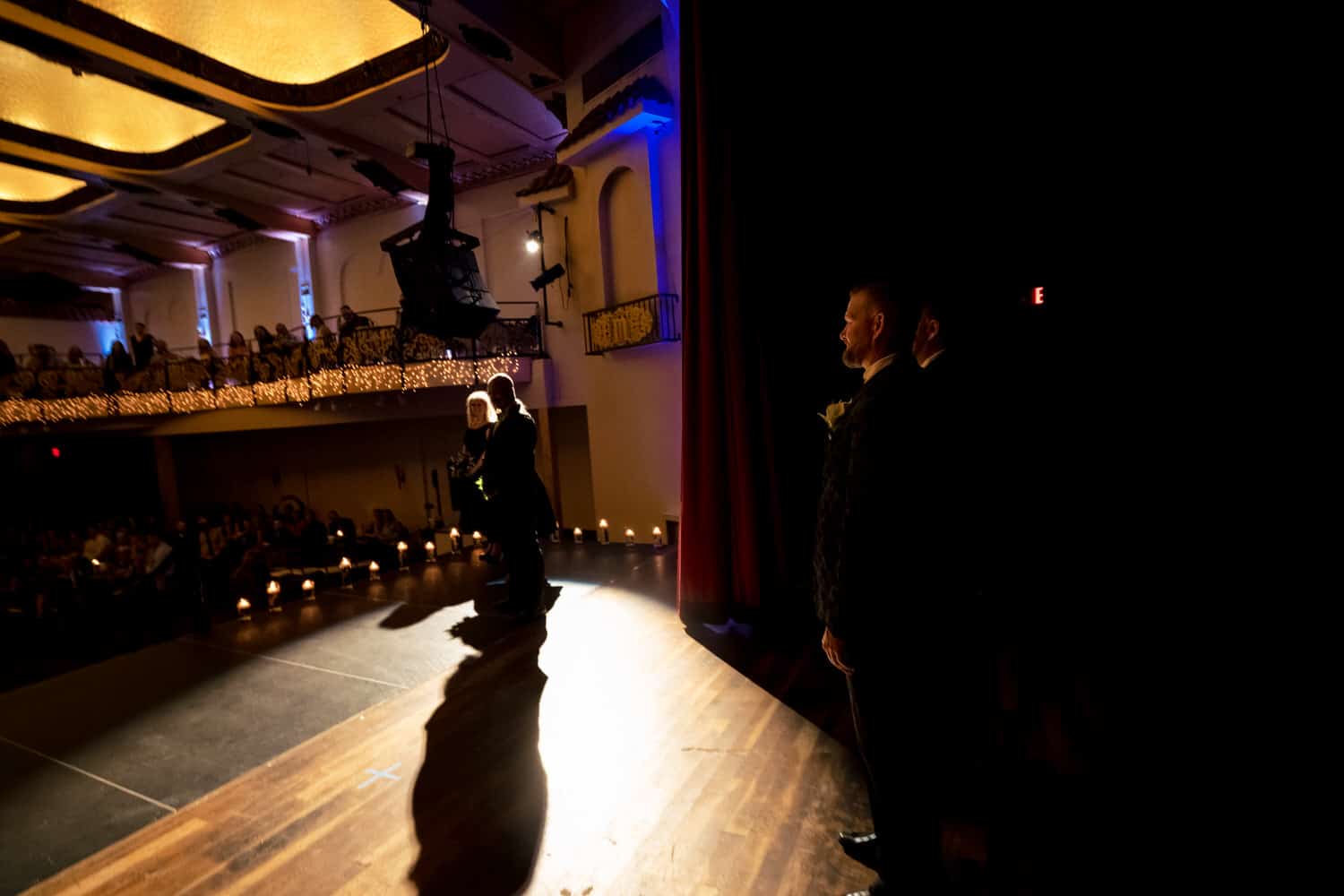 A picture of two grooms holding hands, walking onto the stage of The Madrid Theatre for their wedding ceremony as the curtain rolls back. 