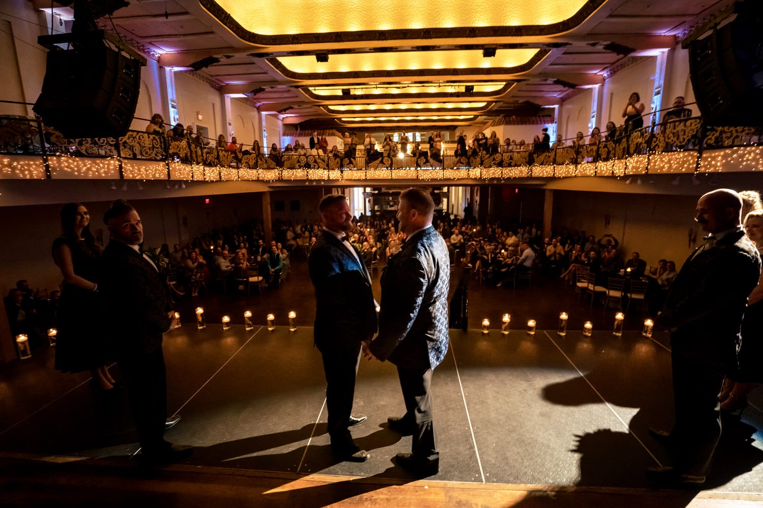A picture taken from the back of the stage looking out towards the audience of two grooms holding hands and exchanging their vows during their wedding ceremony at The Madrid Theatre in Kansas City. 