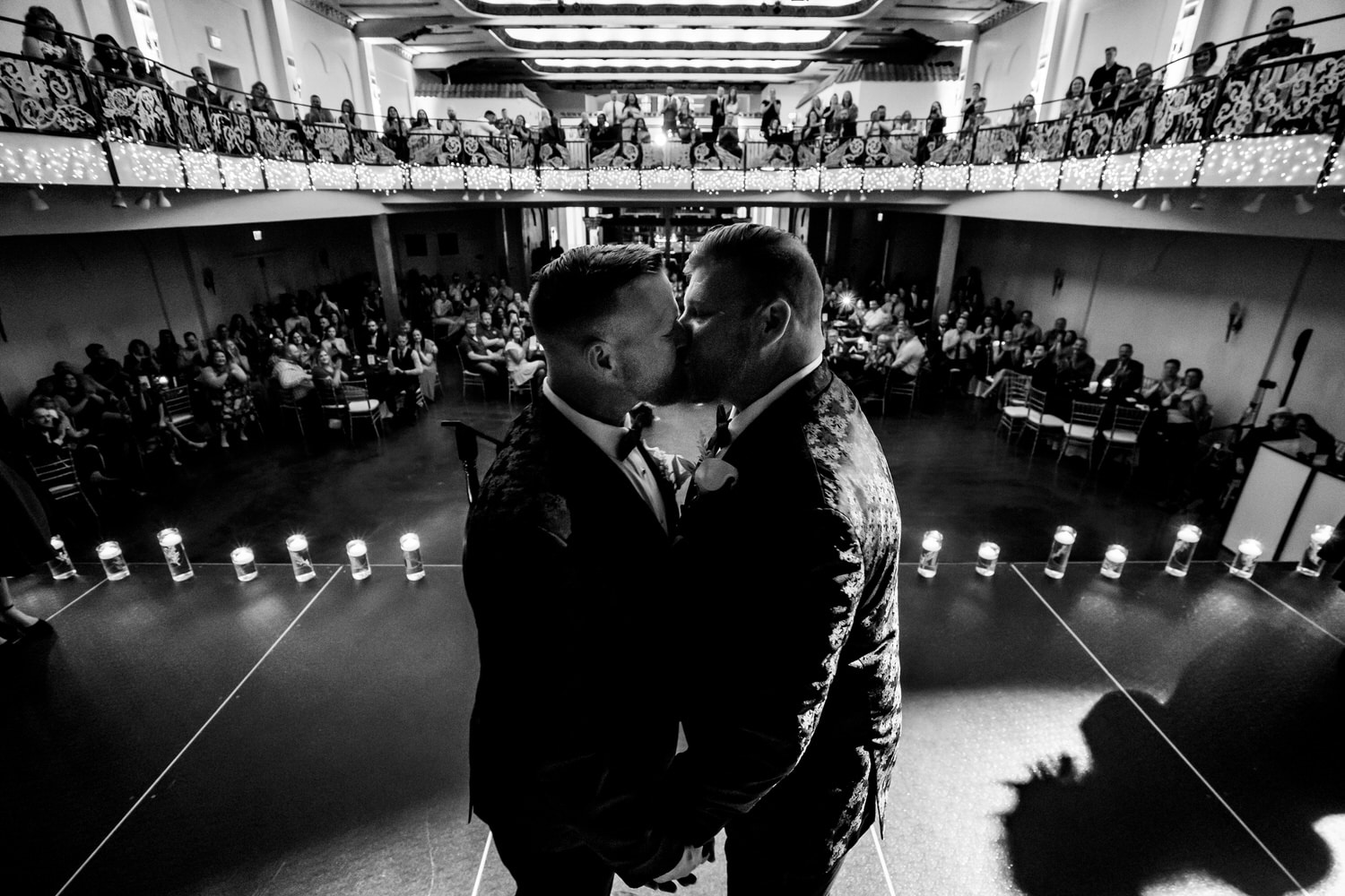 A candid black and white picture of two grooms sharing their first kiss at the end of their fall wedding ceremony at The Madrid Theatre in Kansas City; their guests visible behind them. 