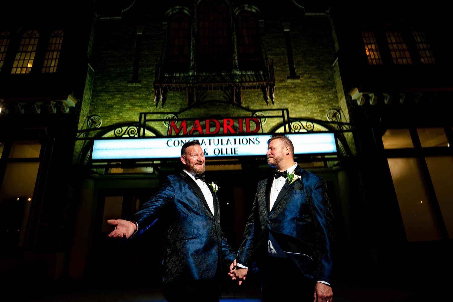 A candid picture of two grooms, holding hands and celebrating in front of The Madrid Theatre on their fall wedding day in Kansas City. 