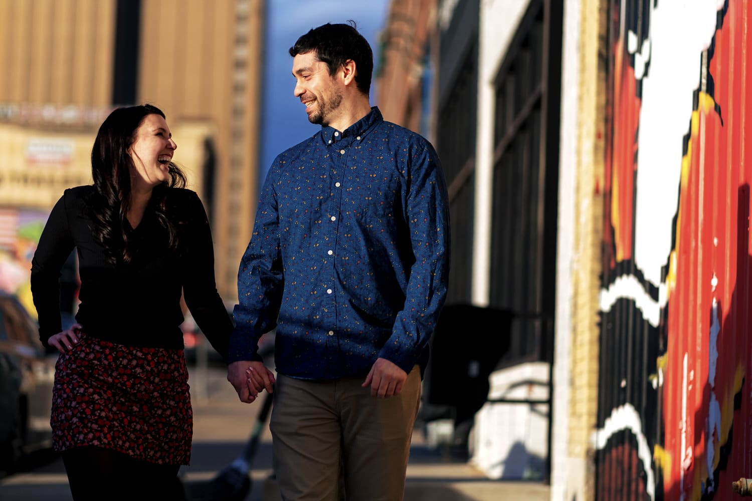 A candid colorful picture of an engaged couple walking down a sidewalk in Kansas City's Crossroads District holding hands and laughing together during their winter engagement session. 