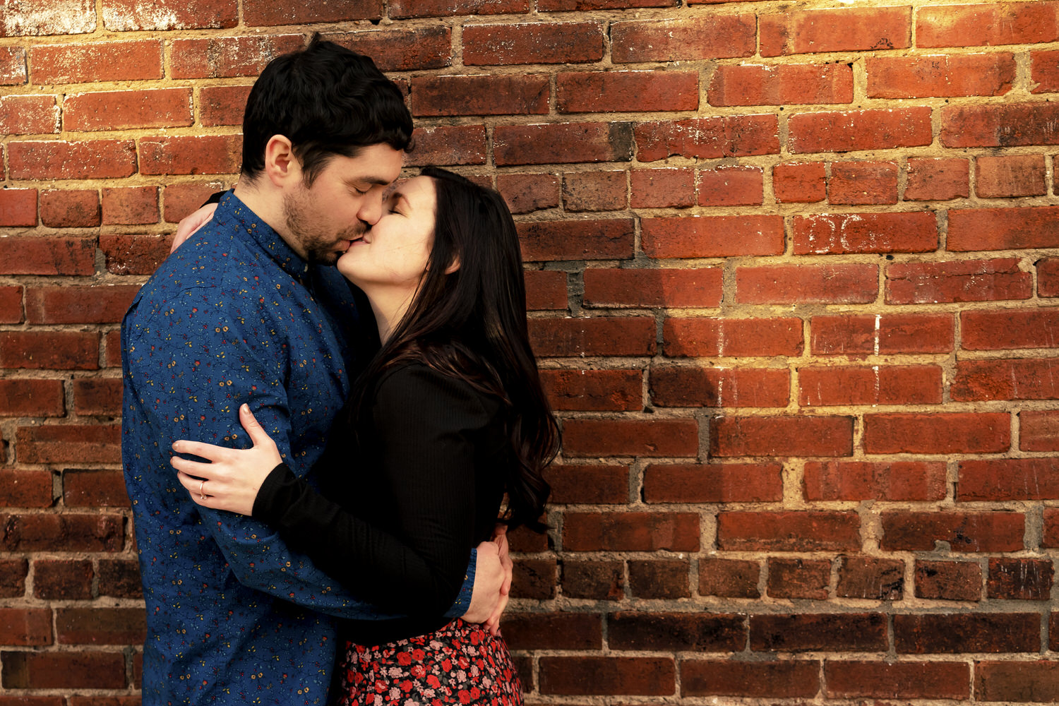 A colorful, candid picture of an engaged couple sharing a kiss in front of a red brick wall during their winter engagement session in Kansas City's Crossroads District. 